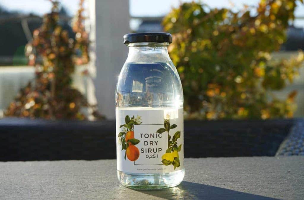 Auf einem Tisch im Freien steht eine durchsichtige Glasflasche mit der Aufschrift „Tonic Dry Sirup 0,25 l“; Sonnenlicht fällt auf die Flasche, während im Hintergrund grünes Laub verschwommen zu sehen ist.