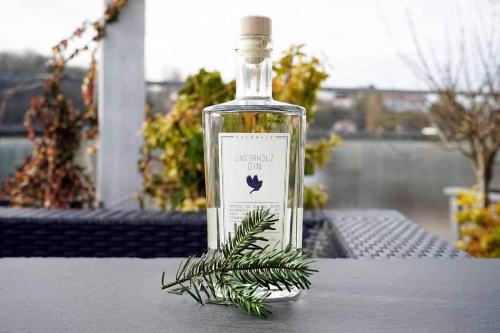 Auf einem Tisch im Freien steht eine durchsichtige Flasche Unterholz Gin mit Korkverschluss neben einem grünen Kiefernzweig. Im Hintergrund sind verschwommene Pflanzen und ein Gewässer zu sehen.