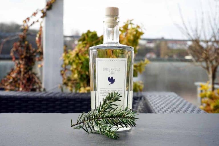 Auf einem Tisch im Freien steht eine durchsichtige Flasche Unterholz Gin mit Korkverschluss neben einem grünen Kiefernzweig. Im Hintergrund sind verschwommene Pflanzen und ein Gewässer zu sehen.
