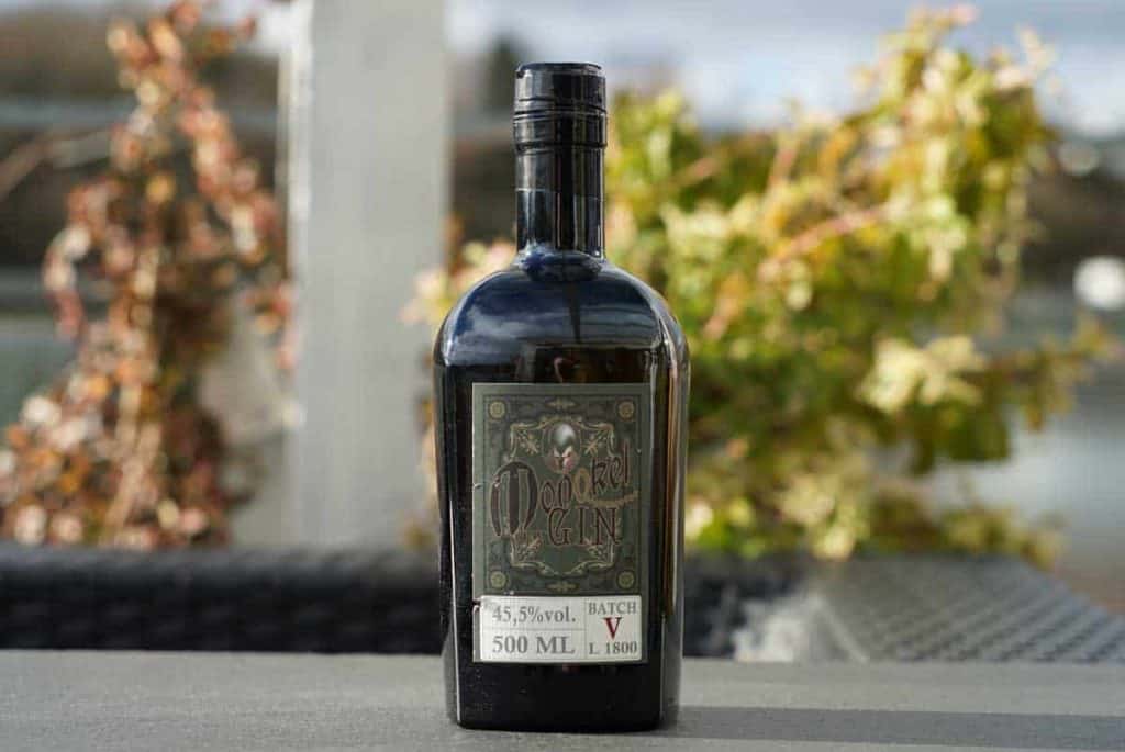 Auf einem Tisch im Freien steht eine dunkle Glasflasche Gin mit der Aufschrift „Opihr Gin“ und einem dekorativen Motiv; im Hintergrund sind verschwommene Pflanzen und ein Geländer zu sehen.
