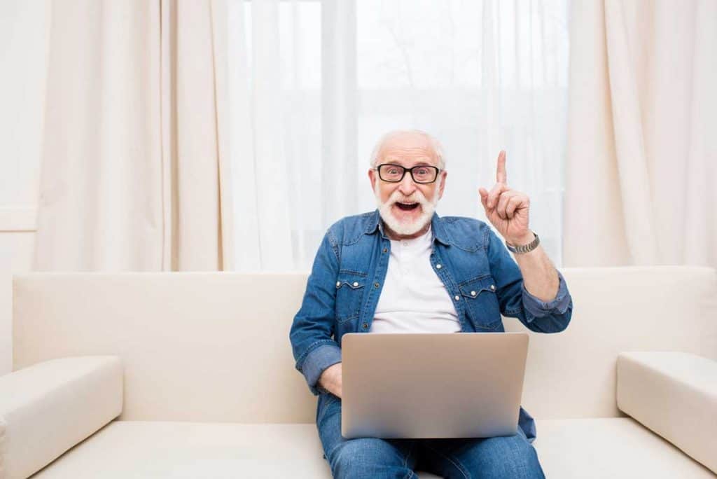 Ein älterer Mann mit Brille sitzt auf einem beigen Sofa und arbeitet an einem Laptop; er lächelt und hebt einen Finger, als wäre ihm gerade eine Idee gekommen. Im Hintergrund sind leichte Vorhänge und ein großes Fenster zu sehen.
