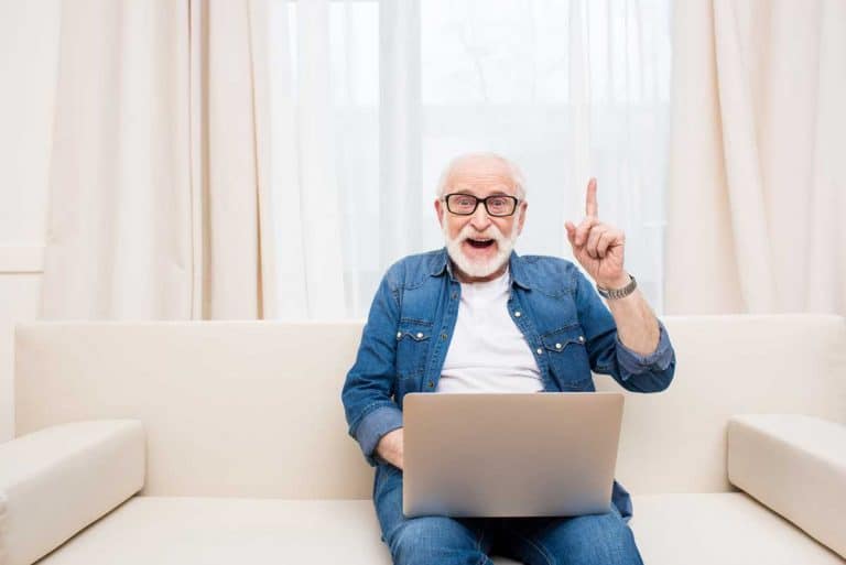 Ein älterer Mann mit Brille sitzt auf einem beigen Sofa und arbeitet an einem Laptop; er lächelt und hebt einen Finger, als wäre ihm gerade eine Idee gekommen. Im Hintergrund sind leichte Vorhänge und ein großes Fenster zu sehen.