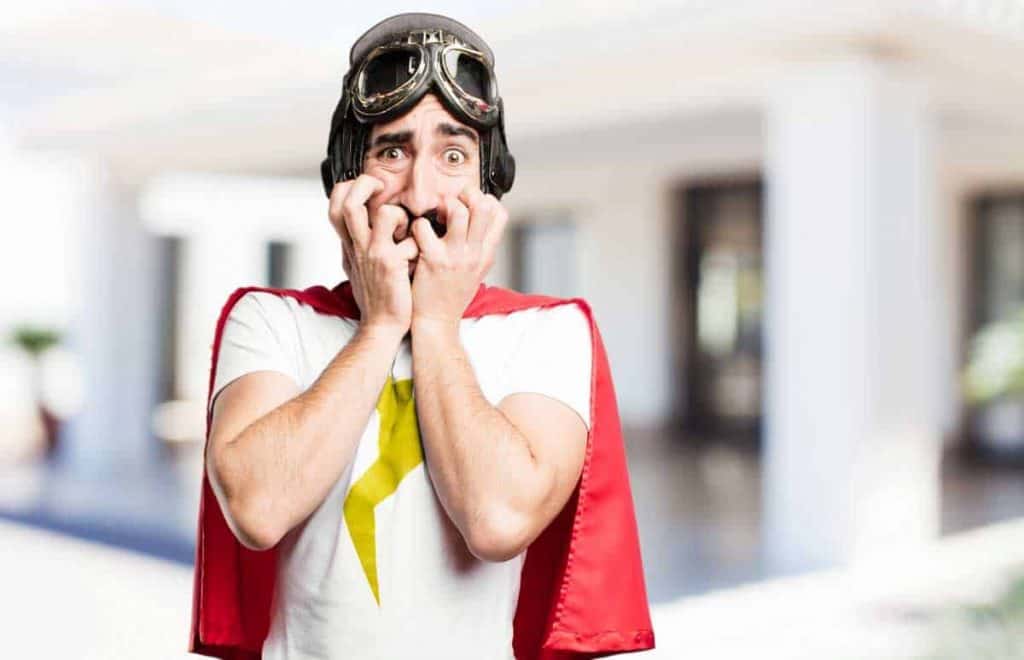 Ein Mann in einem roten Umhang und einer Pilotenbrille wirkt nervös, beißt sich auf die Finger und starrt mit weit aufgerissenen Augen in einen hellen, verschwommenen Innenraum.