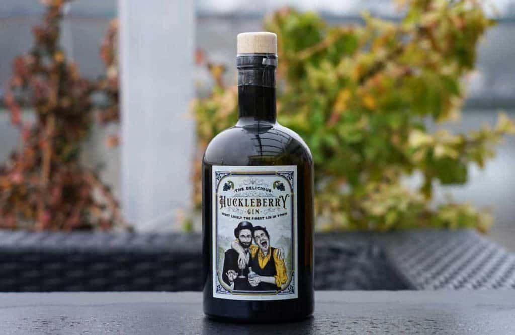 Eine dunkle Glasflasche mit Huckleberry Gin und Korkverschluss, versehen mit einem illustrierten Etikett, auf dem zwei Männer zu sehen sind; die Flasche steht im Freien auf einem Tisch, im Hintergrund sind verschwommene Pflanzen und ein Geländer zu erkennen.