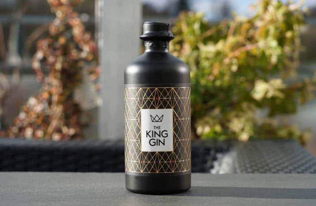 Auf einem Tisch im Freien steht eine schwarze Flasche mit der Aufschrift „The King Gin“ und einem goldenen geometrischen Muster; im Hintergrund sind verschwommene Pflanzen und ein Geländer zu sehen.