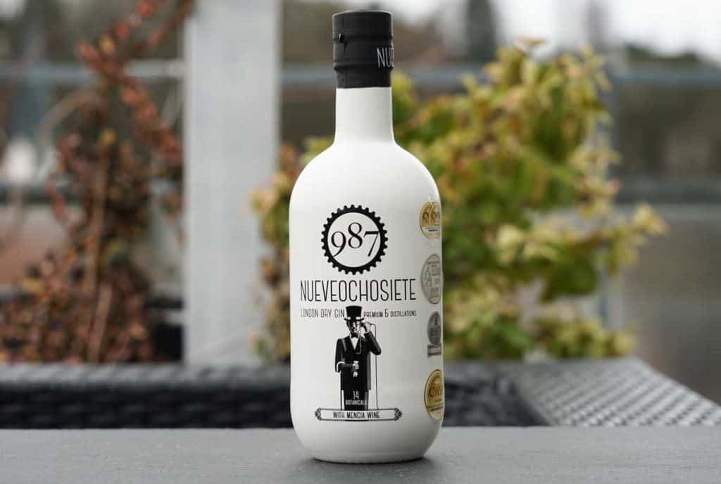 Auf einem Tisch im Freien steht eine Flasche „987 NUEVEOCHOSIETE London Dry Gin“, im Hintergrund sind verschwommen Grünflächen und Gebäude zu sehen.