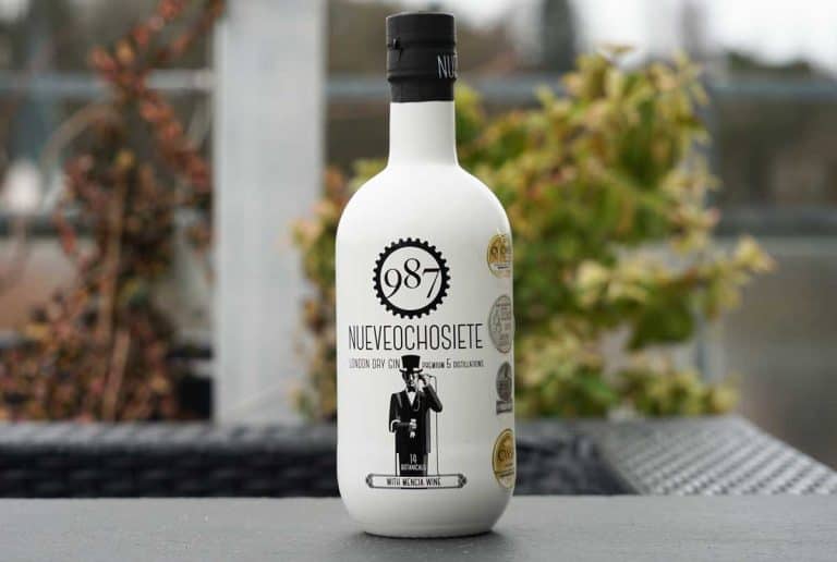 Auf einem Tisch im Freien steht eine Flasche „987 NUEVEOCHOSIETE London Dry Gin“, im Hintergrund sind verschwommen Grünflächen und Gebäude zu sehen.