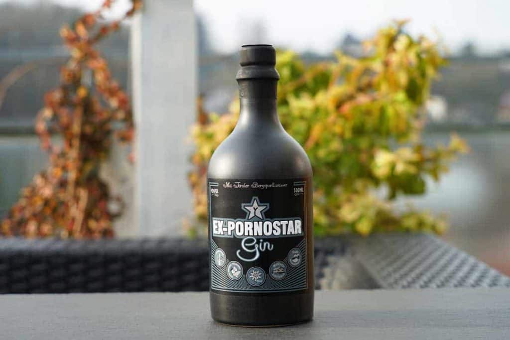 Auf einem dunklen Tisch im Freien steht eine schwarze Flasche mit der Aufschrift „Ex-Pornostar Gin“, im Hintergrund sind grünes Laub sowie ein verschwommener Fluss und eine Brücke zu sehen.