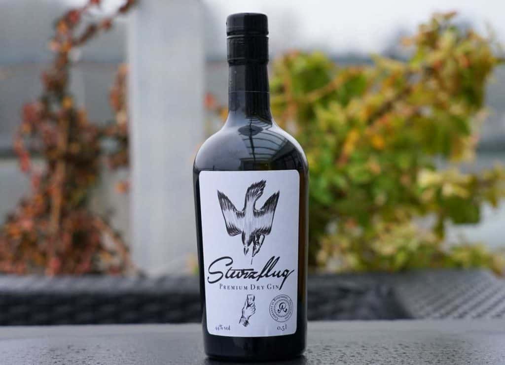 Eine Flasche „Sturzflug Premium Dry Gin“ mit schwarzem Verschluss und weißem Etikett, auf dem ein fliegender Vogel abgebildet ist, im Freien vor einem unscharfen Grünhintergrund.