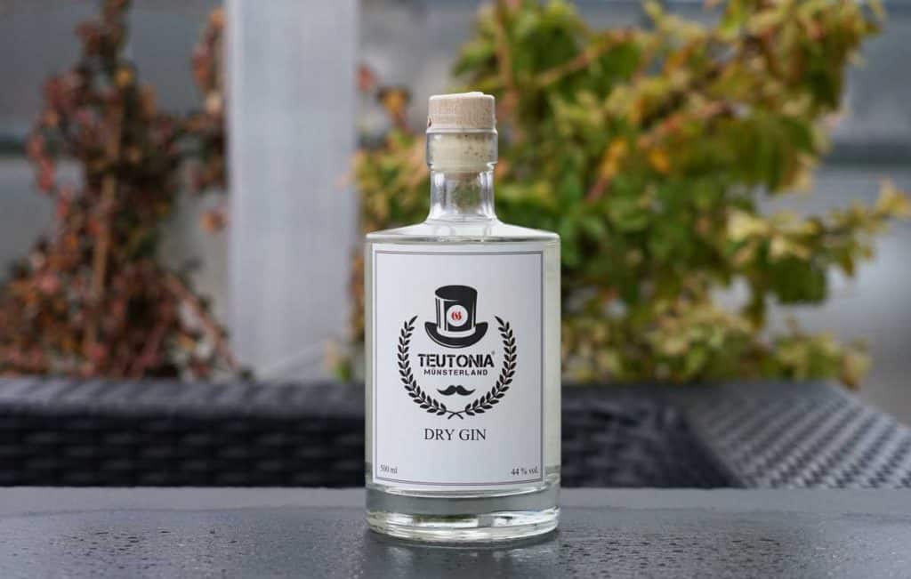 Auf einem dunklen Tisch im Freien steht eine Flasche „Teutonia Münsterland Dry Gin“ mit Korkverschluss, im Hintergrund ist grünes, verschwommenes Laub zu sehen.
