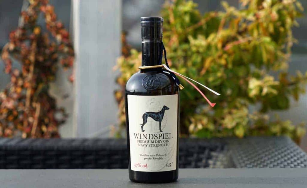 Auf einem Tisch im Freien steht eine Flasche „Windspiel Premium Dry Gin“, im Hintergrund ist verschwommenes grünes und braunes Laub zu sehen. Auf der Flasche befindet sich ein Etikett mit der Abbildung eines schwarzen Hundes.
