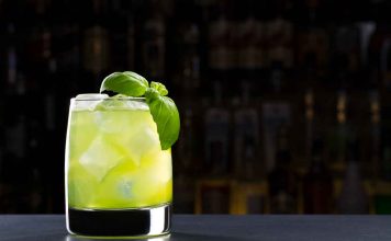 Gin Basil Smash – Gin Cocktail Ein Glas mit einem grünen Cocktail auf Eis, garniert mit einem Zweig frischem Basilikum, steht auf einer dunklen Fläche vor einem unscharfen Barhintergrund.