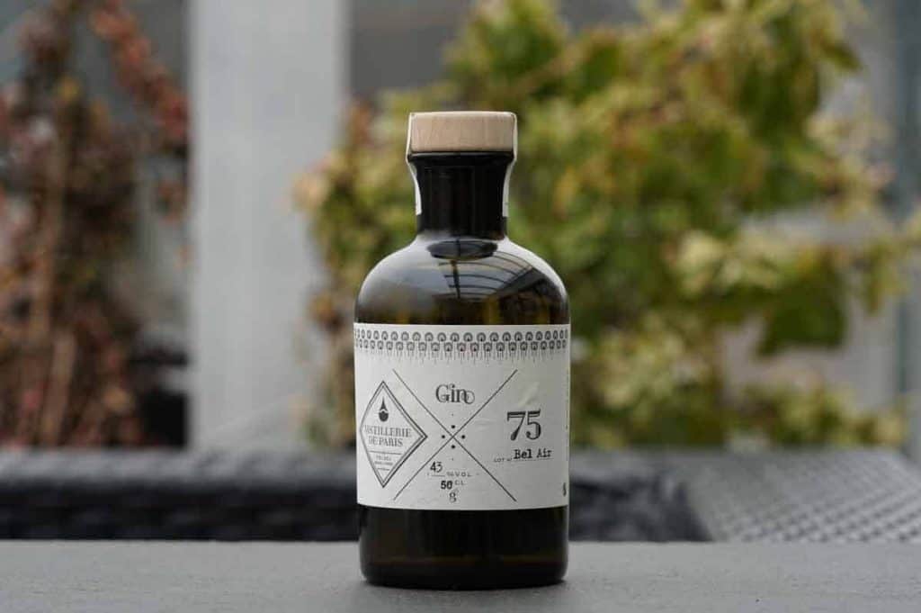 Eine dunkle Glasflasche mit einem beigen Verschluss und einem weißen Etikett mit geometrischen Mustern und Text, die auf einer grauen Fläche steht, im Hintergrund ist verschwommenes Grün zu sehen.