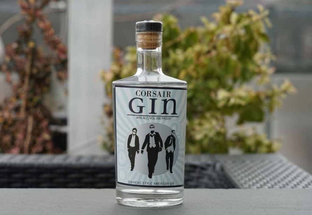 Eine durchsichtige Flasche Corsair Gin mit einem Etikett, auf dem drei Personen in Anzügen und mit Sonnenbrillen zu sehen sind, die im Freien auf einer grauen Fläche stehen, im Hintergrund sind verschwommene grüne Pflanzen zu erkennen.