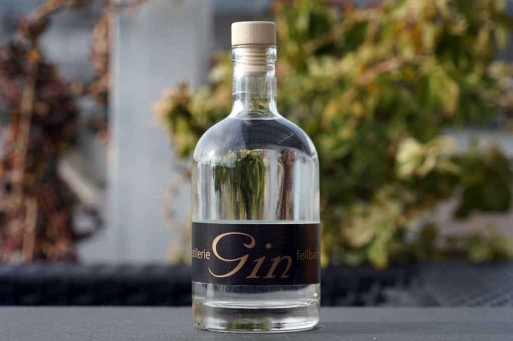 Auf einer Fläche steht eine Gin-Flasche aus klarem Glas mit einem Holzverschluss und einem schwarzen Etikett, im Hintergrund ist verschwommenes grünes und braunes Laub zu sehen.