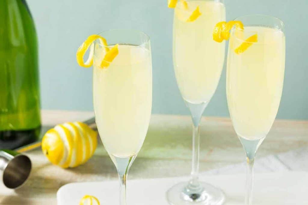Drei Champagnergläser, gefüllt mit einem hellgelben Cocktail und mit Zitronenspiralen garniert. Im Hintergrund sind auf einer hellen Unterlage eine grüne Flasche, ein Jigger und eine Zitrone zu sehen.