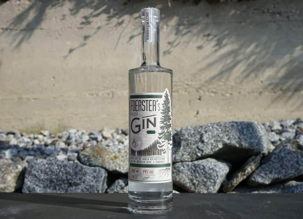 Eine Flasche Foersters Heide Gin steht im Freien auf einer dunklen Fläche, im Hintergrund sind Felsen und eine Steinmauer zu sehen. Auf dem Etikett sind Bäume und grüne Akzente abgebildet.