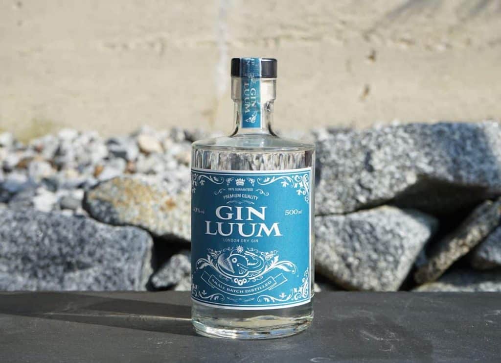 Eine Flasche „Gin Luum London Dry Gin“ mit blauem Etikett steht auf einer dunklen Fläche vor grauen Felsen und einer beigen Wand.