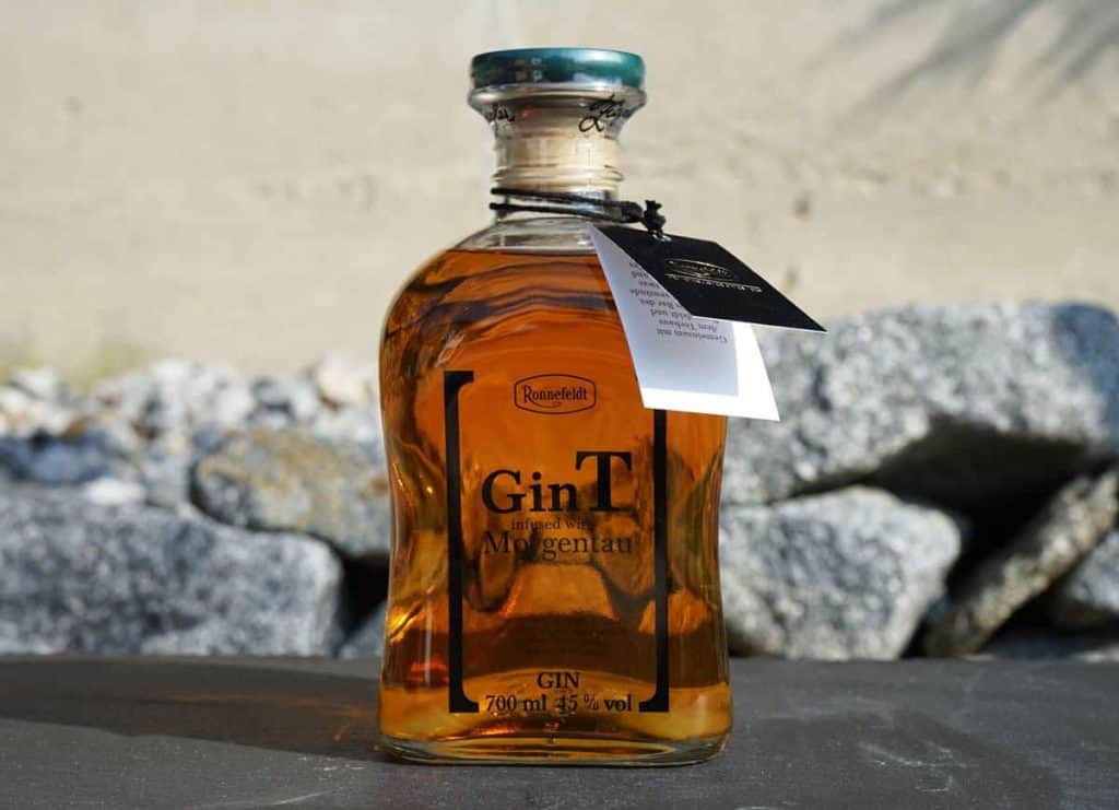 Eine Flasche goldfarbener Gin mit der Aufschrift „GinT“ und einem schwarzen Etikett um den Flaschenhals steht auf einer Fläche mit Steinen, im Hintergrund ist eine beigefarbene Wand zu sehen.