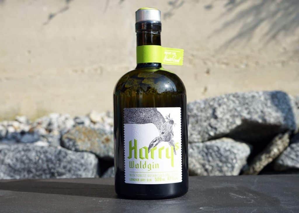 Eine Flasche Harry Waldgin London Dry Gin steht auf einer dunklen Fläche, im Hintergrund sind Felsen und eine beigefarbene Wand zu sehen. Das Etikett zeigt ein Waldmotiv, und am Flaschenhals befindet sich ein grünes Etikett.