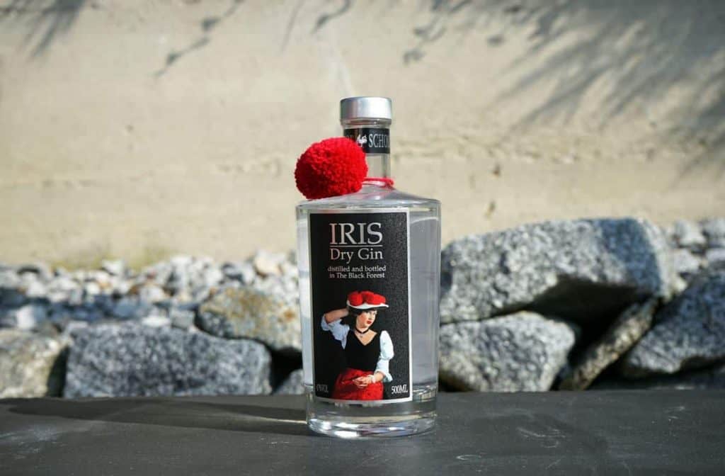 Eine Flasche IRIS Dry Gin mit einem roten Pompon am Flaschenhals steht im Freien auf einer dunklen Fläche, im Hintergrund sind Felsen und eine Betonmauer zu sehen. Auf dem Etikett ist eine Frau in traditioneller Kleidung abgebildet.