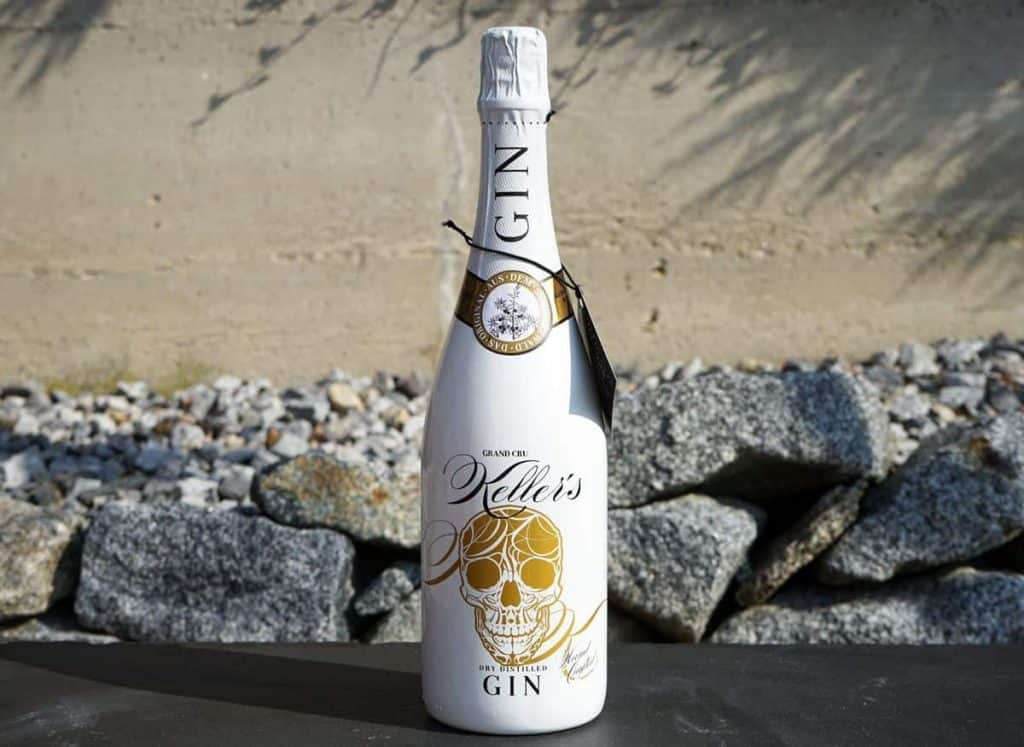 Auf einer schwarzen Fläche steht eine weiße Gin-Flasche mit einem goldenen Totenkopf-Motiv und dem Etikett „Vetters Dry Distilled Gin“, im Hintergrund sind Felsen und eine beige Wand zu sehen.