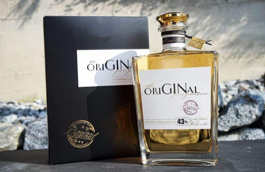 Eine Flasche „The Original“-Schnaps in einer schwarz-goldenen Schachtel, die im Freien auf einer Steinfläche steht, vor einem unscharfen Hintergrund aus Steinen und einer Mauer.