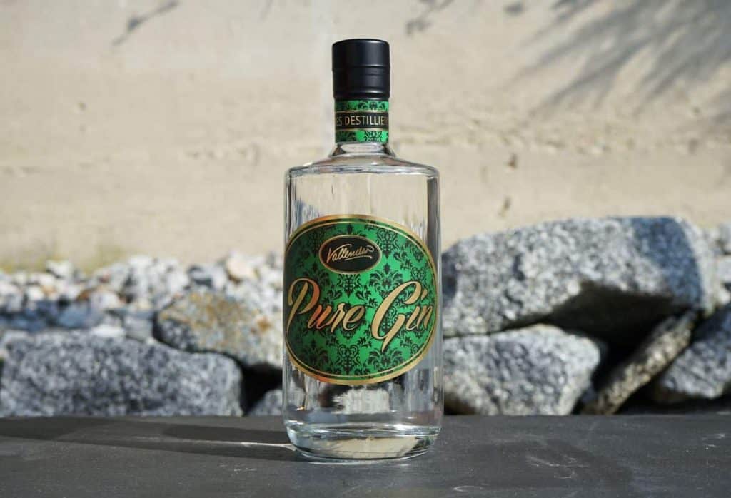 Auf einer dunklen Fläche steht eine durchsichtige Glasflasche mit der Aufschrift „Pure Gin“ und einem grün-goldenen Design, im Hintergrund sind graue Felsen und eine beige Wand zu sehen.