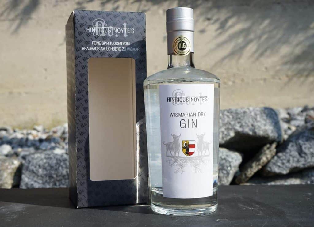Eine Flasche „Hintzious Noytes Wismarian Dry Gin“ steht neben ihrer grau-weißen Schachtel; beide Gegenstände sind auf einer Steinoberfläche vor einem felsigen Hintergrund platziert.