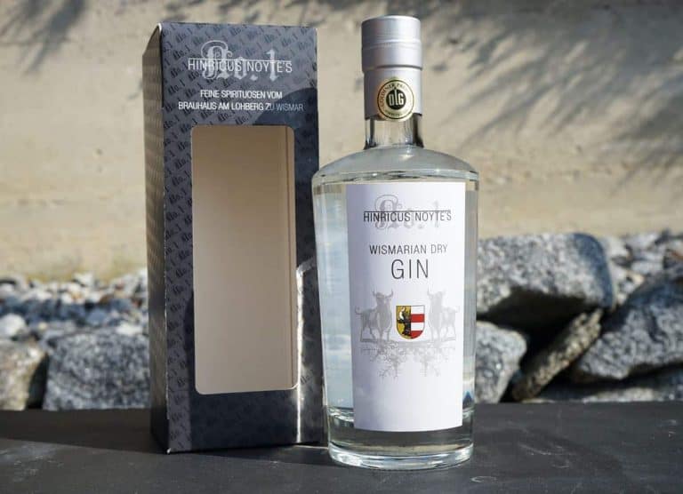 Eine Flasche „Hintzious Noytes Wismarian Dry Gin“ steht neben ihrer grau-weißen Schachtel; beide Gegenstände sind auf einer Steinoberfläche vor einem felsigen Hintergrund platziert.