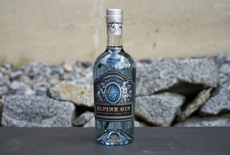 Auf einer dunklen Fläche steht eine Flasche Alpine Gin mit silbernem Verschluss und verziertem Etikett, im Hintergrund sind graue Steine und eine beige Wand zu sehen.