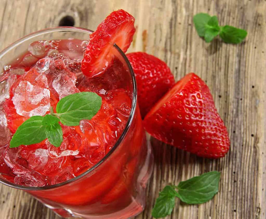 Ein Glas Erdbeer-Eisgetränk, garniert mit einem Minzblatt und einer Erdbeerscheibe, dazu frische Erdbeeren und Minzblätter auf einer rustikalen Holzunterlage.