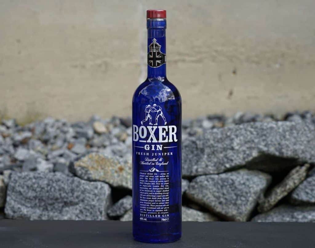 Eine blaue Flasche Boxer Gin mit rotem Verschluss steht auf einer dunklen Fläche, im Hintergrund sind graue Felsen und eine beige Wand zu sehen.
