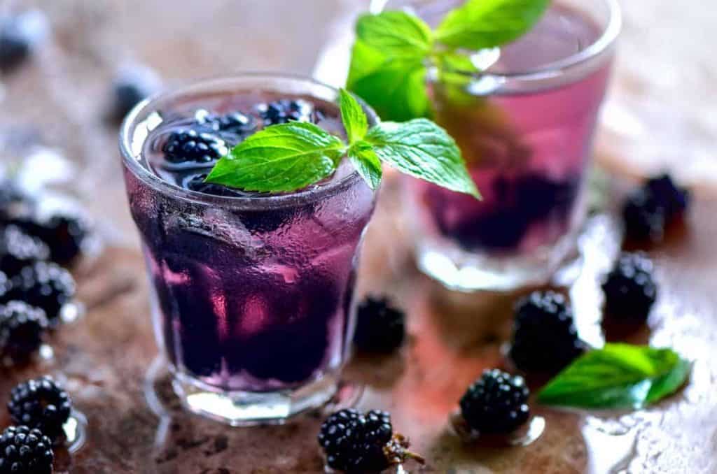 Zwei Gläser mit violettem Brombeergetränk auf Eis, garniert mit frischen Minzblättern. Brombeeren und Minzblätter sind auf einer feuchten braunen Fläche rund um die Gläser verstreut.