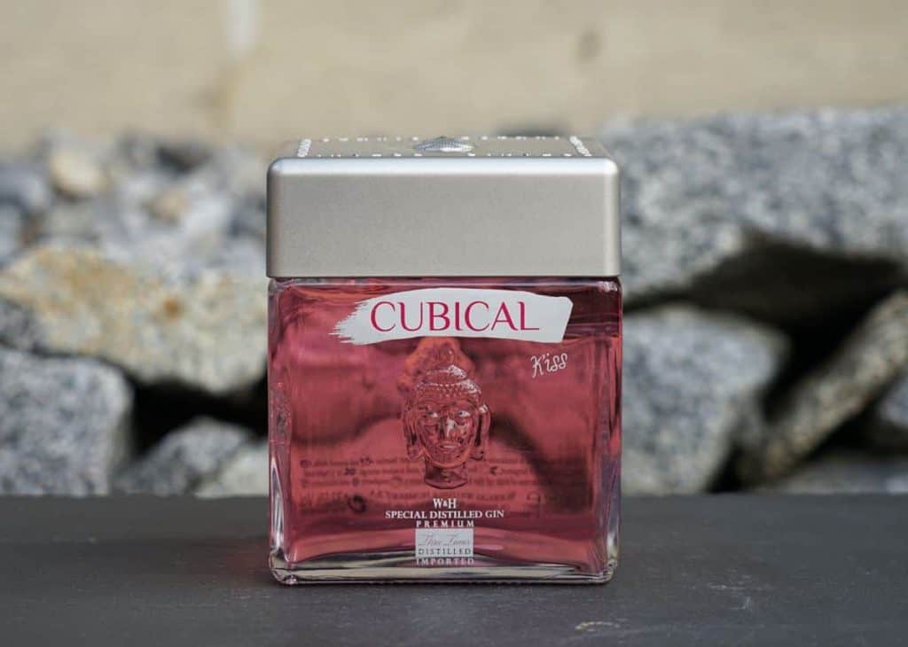 Eine quadratische Glasflasche mit „Cubical“-Gin und einem silbernen Verschluss, die eine rosafarbene Flüssigkeit enthält, steht auf einer dunklen Oberfläche vor einem Hintergrund aus grauen Felsen.