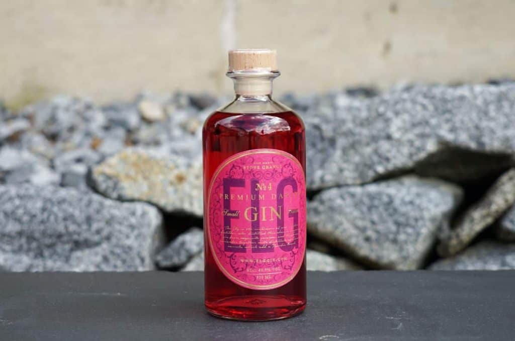 Eine durchsichtige Flasche mit rosa Gin, einem Holzkorken und einem leuchtend rosa Etikett steht auf einer dunklen Fläche, im Hintergrund sind graue Steine verschwommen zu erkennen.