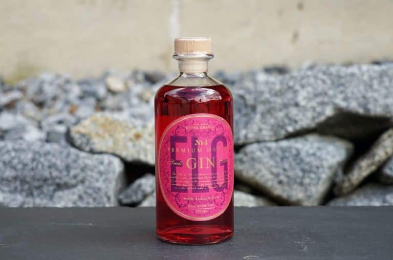 Eine durchsichtige Flasche mit rosa Gin, einem Holzkorken und einem leuchtend rosa Etikett steht auf einer dunklen Fläche, im Hintergrund sind graue Steine verschwommen zu erkennen.