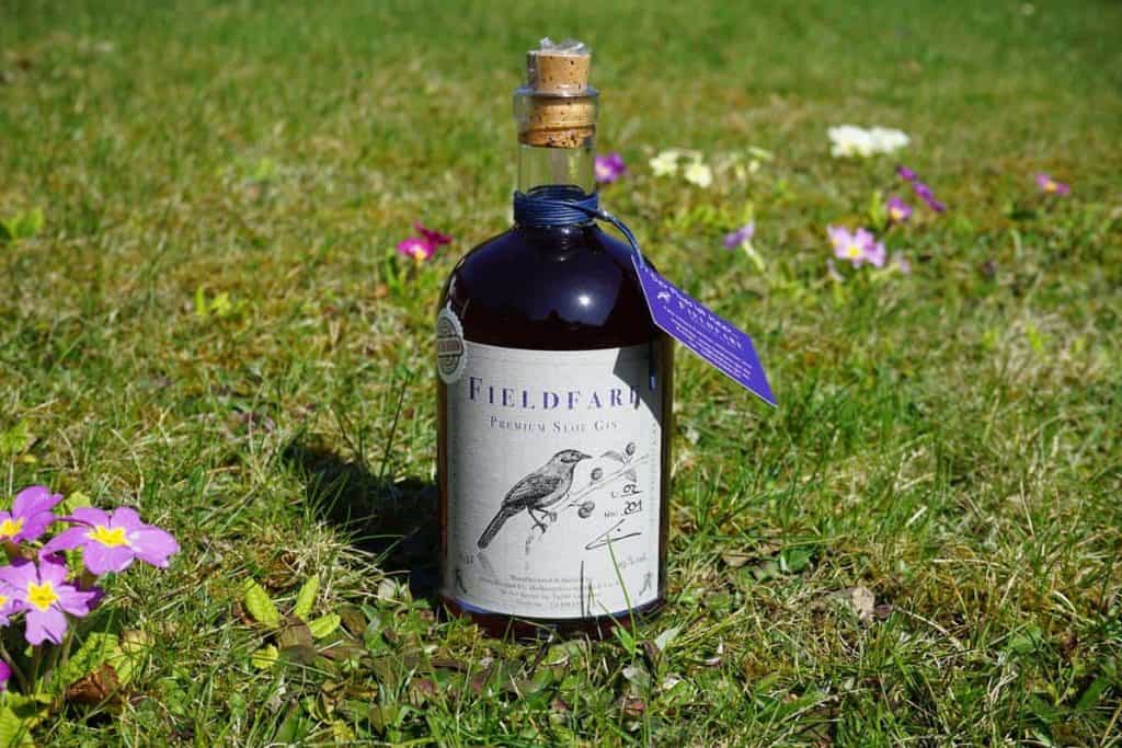 Eine Flasche „Fieldfare“-Gin mit einer Vogelabbildung auf dem Etikett steht an einem sonnigen Tag auf einer Wiese, umgeben von kleinen violetten und weißen Blüten.
