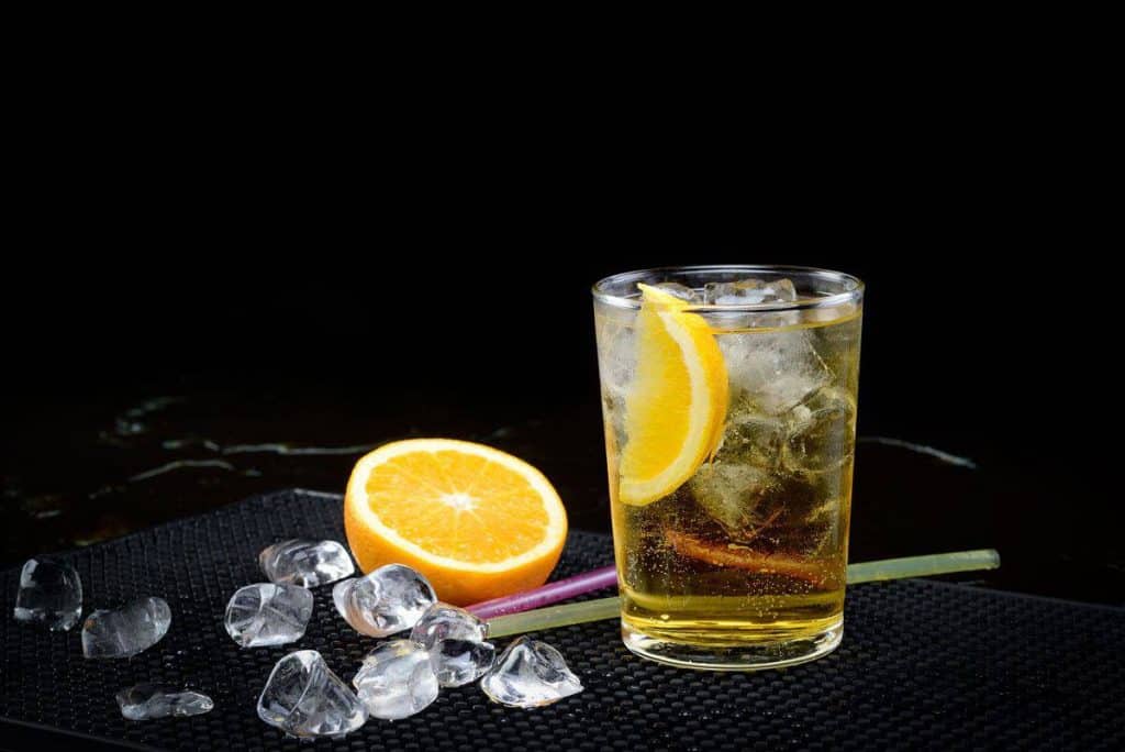 Ein Glas mit einem gekühlten Getränk, garniert mit einer Zitronenscheibe, steht neben einer halbierten Orange und verstreuten Eiswürfeln auf einer schwarzen Fläche vor einem dunklen Hintergrund.