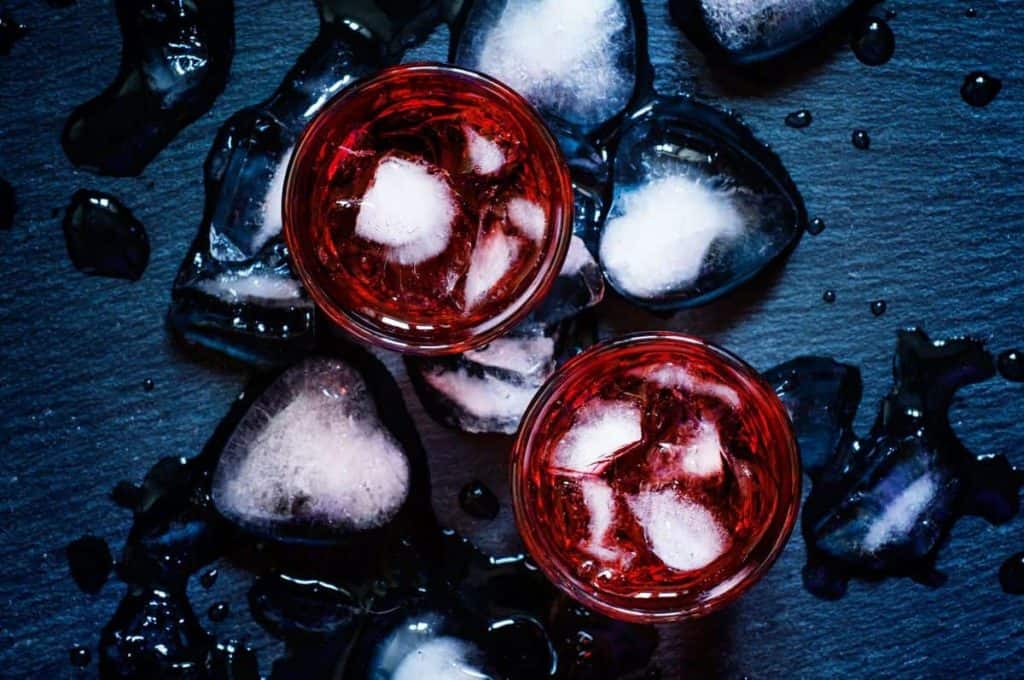 Zwei Gläser, gefüllt mit einem roten Getränk und Eiswürfeln, stehen auf einer dunklen Fläche, auf der geschmolzene Eiswürfel und Wassertropfen verstreut sind.