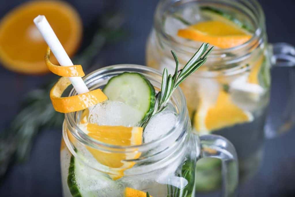 Zwei Einmachgläser, gefüllt mit Eis, Gurkenscheiben, Orangenschnitzen und Rosmarinzweigen, mit einem Papierstrohhalm, auf dem eine Orangenschalenspirale liegt. Ein erfrischendes aromatisiertes Wassergetränk.