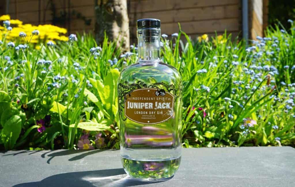 Eine durchsichtige Flasche „Juniper Jack London Dry Gin“ steht im Freien auf einer dunklen Fläche, umgeben von grünem Gras, blauen Blumen und Sonnenlicht.