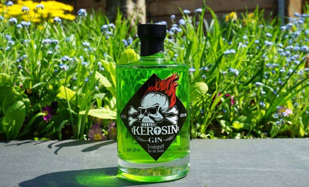 Eine grüne Flasche „Kerosin Gin“ mit einem Etikett, auf dem ein Totenkopf und Flammen abgebildet sind, steht auf einer Steinfläche, im Hintergrund sind grünes Gras und blaue Blumen zu sehen.
