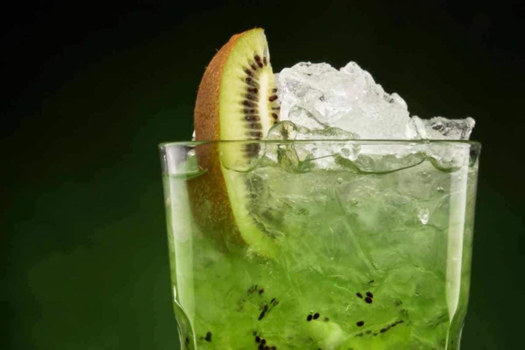 Eine Nahaufnahme eines grünen Kiwi-Cocktails in einem mit zerstoßenem Eis gefüllten Glas, garniert mit einer Scheibe frischer Kiwi, vor einem dunkelgrünen Hintergrund.