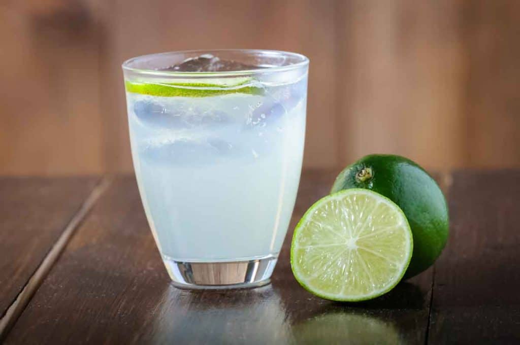 Auf einem Holztisch steht ein Glas Limonade mit Eis und Limettenscheiben neben einer ganzen Limette und einer halben Limette.