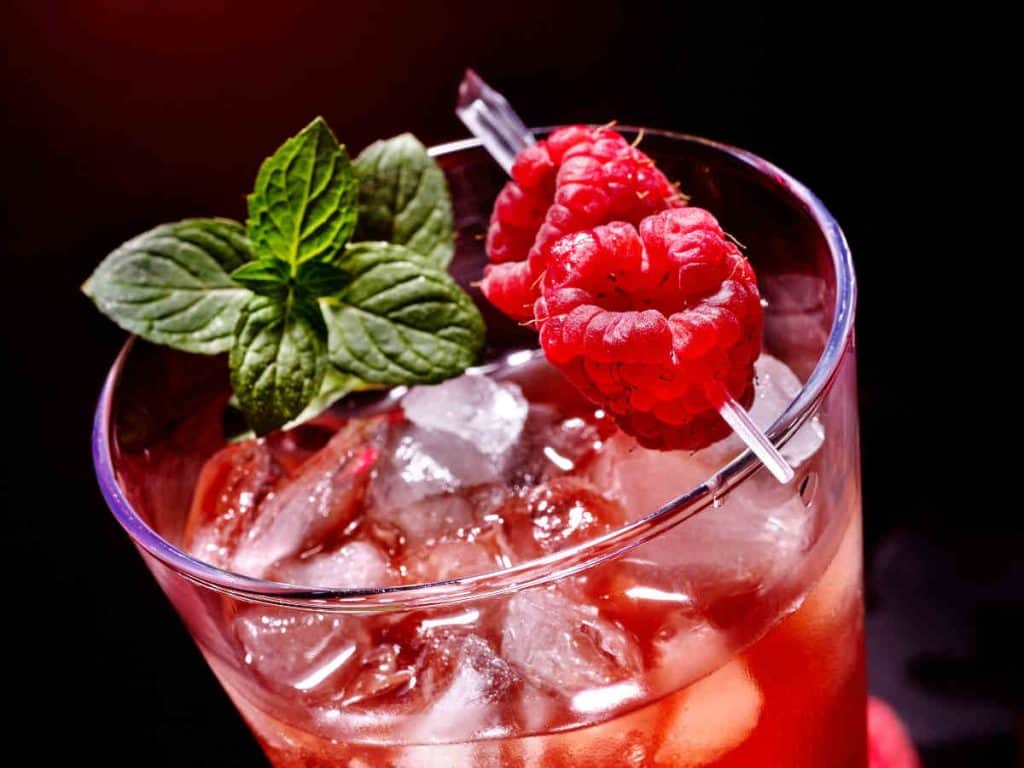Ein Glas Himbeer-Eis-Cocktail, garniert mit zwei frischen Himbeeren am Spieß und einem Minzzweig, vor einem dunklen Hintergrund.
