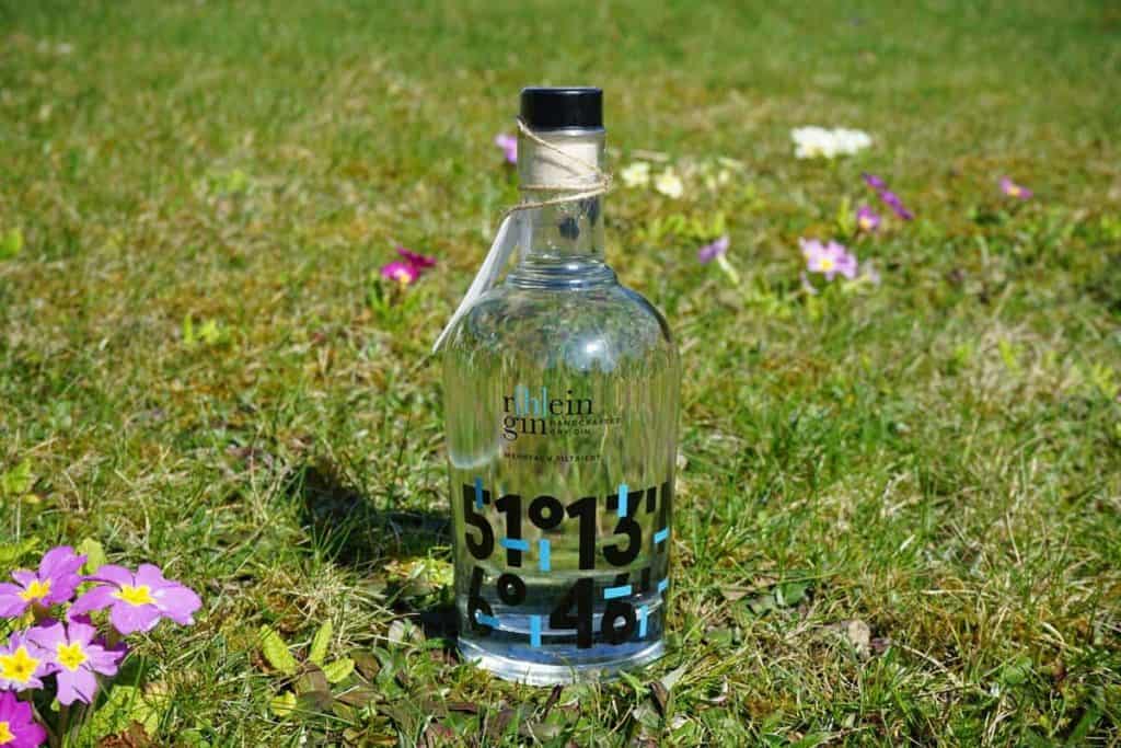 Eine Gin-Flasche aus Glas mit schwarzem Aufdruck steht auf grünem Gras, umgeben von violetten, gelben und weißen Blumen, im natürlichen Sonnenlicht.