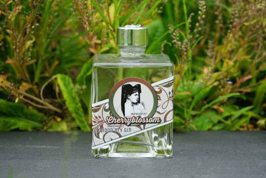 Eine quadratische Glasflasche „Cherryblossom London Dry Gin“ mit einem Etikett im Vintage-Stil, auf dem das Porträt einer Frau zu sehen ist, im Freien vor dem Hintergrund grüner Pflanzen.