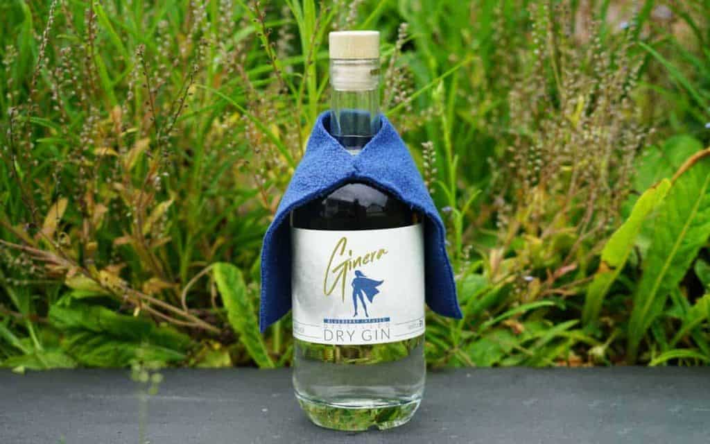 Eine Flasche trockener Gin, über deren „Schultern“ wie ein Umhang ein blaues Tuch drapiert ist, steht im Freien vor grünen Pflanzen.