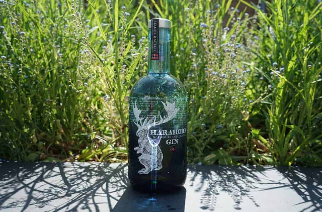Eine Flasche Harahorn-Gin steht im Freien auf einer schwarzen Fläche, umgeben von hohem grünem Gras und Wildblumen im strahlenden Sonnenlicht.